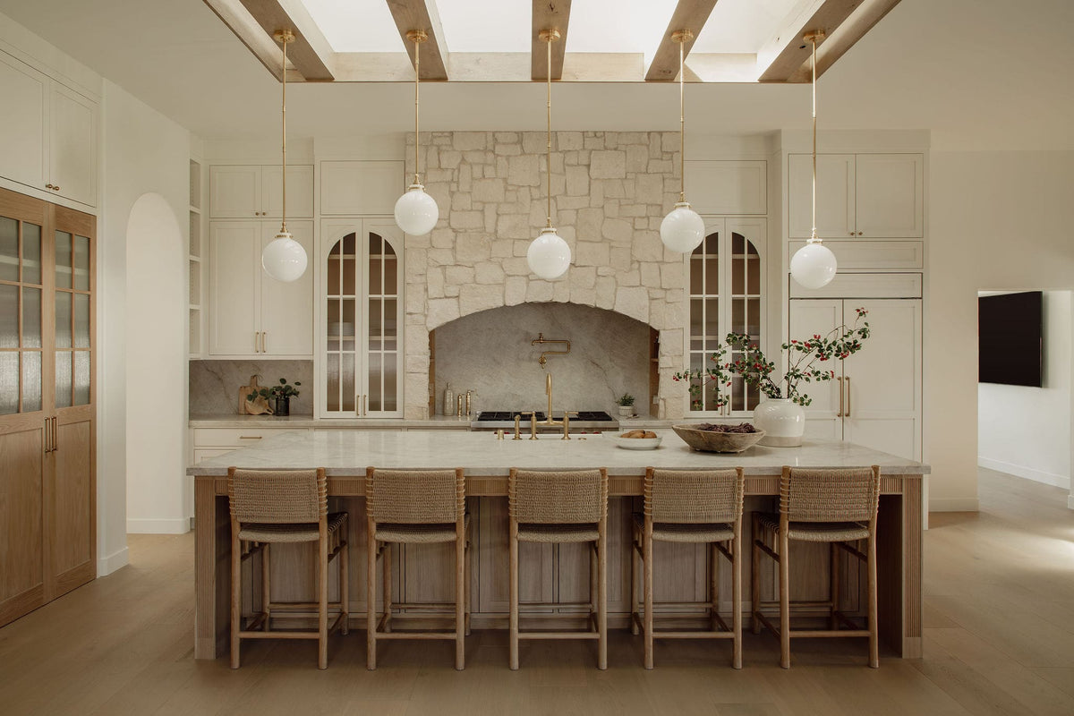 Custom kitchen by Joey Snow with stone arch range hood, white cabinetry, brass fixtures, globe pendant lights, and large wood island.