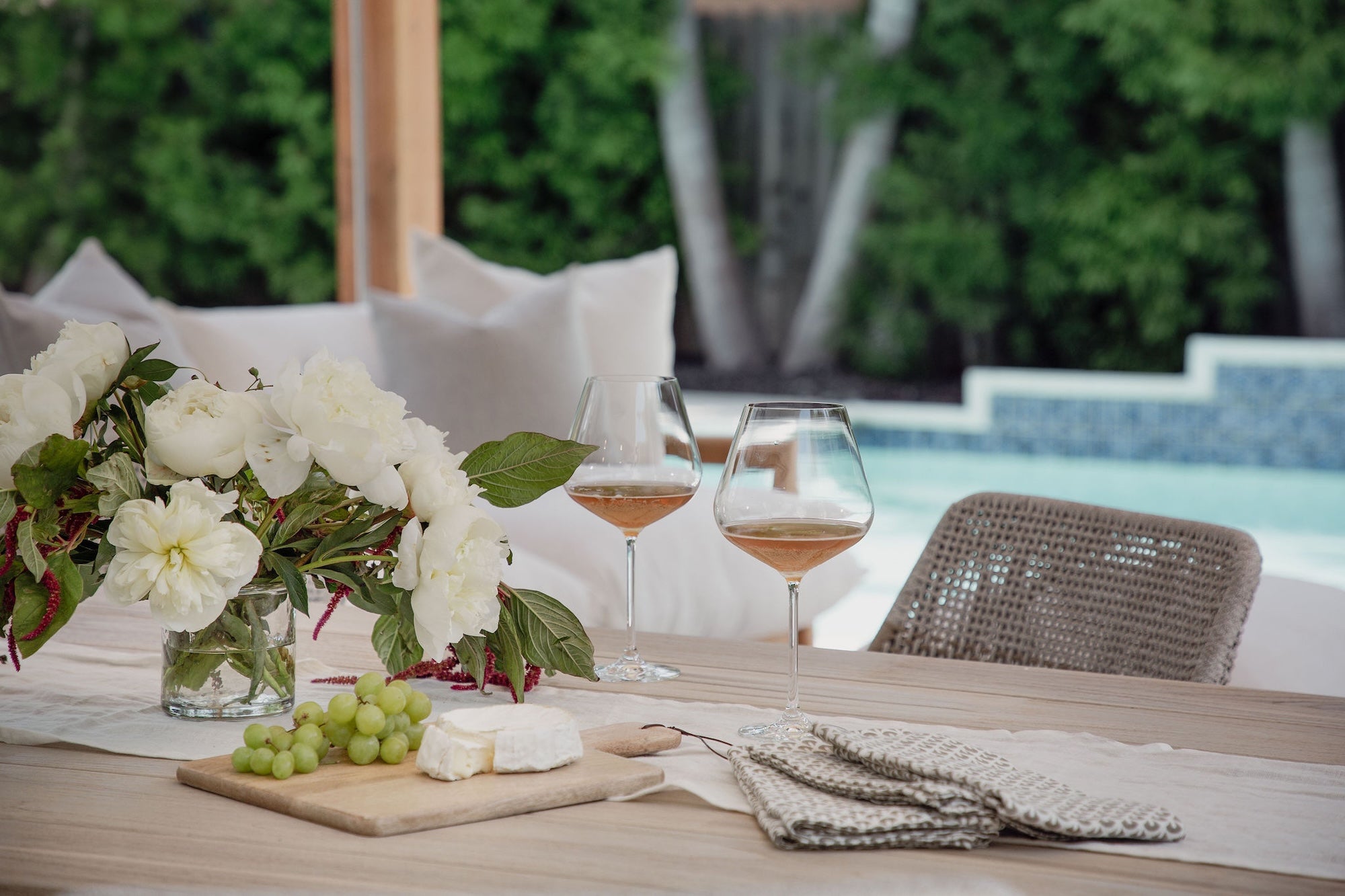 Outdoor dining setup with wine glasses, cheese, grapes, and flowers on a table by a pool.