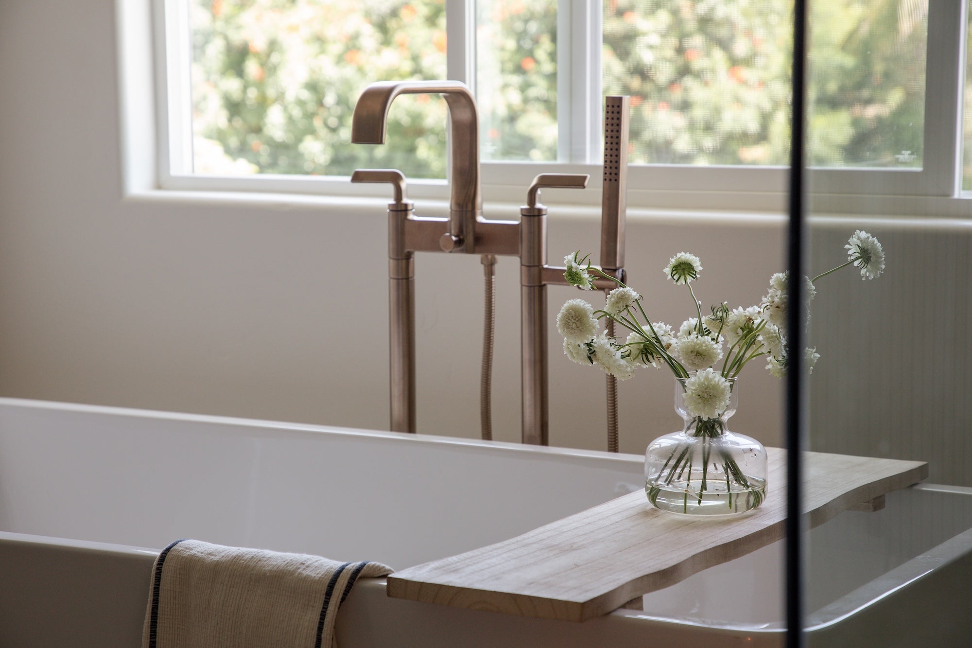 Bathroom setting with a bathtub, wooden tray, and vase of flowers near a window.