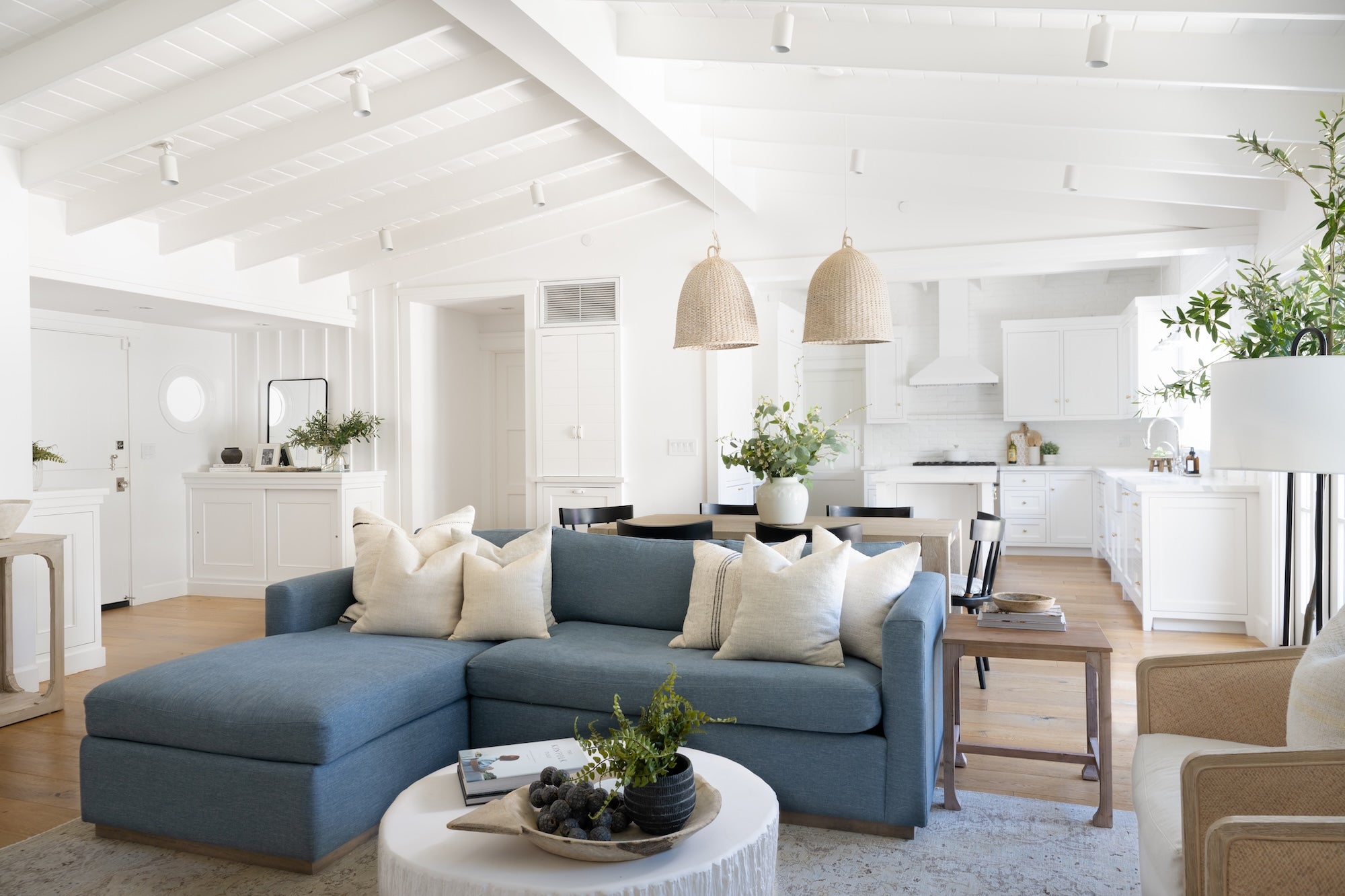 Open-concept living room and kitchen with blue sectional, white beams, and woven pendants.