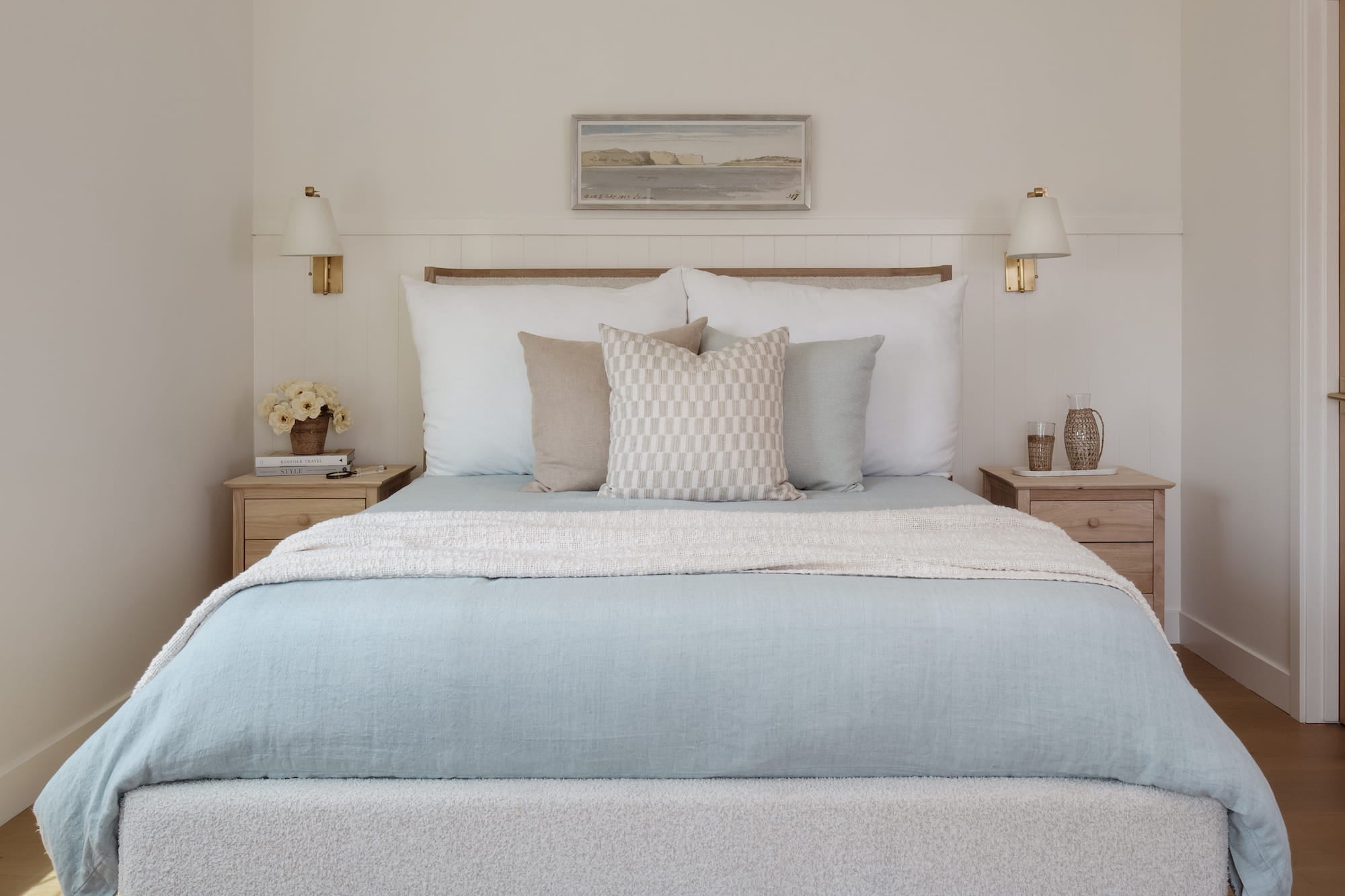 Bedroom with paneled headboard wall, brass sconces, wood nightstands, and layered blue bedding.