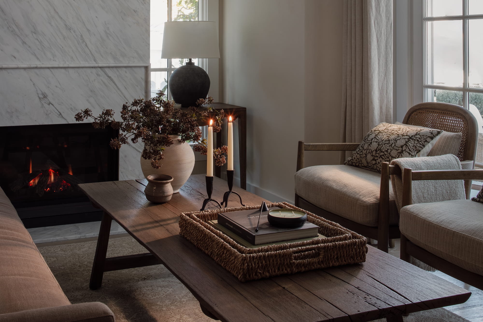 Neutral sofa with patterned pillows beside marble fireplace and styled wood coffee table with woven tray.