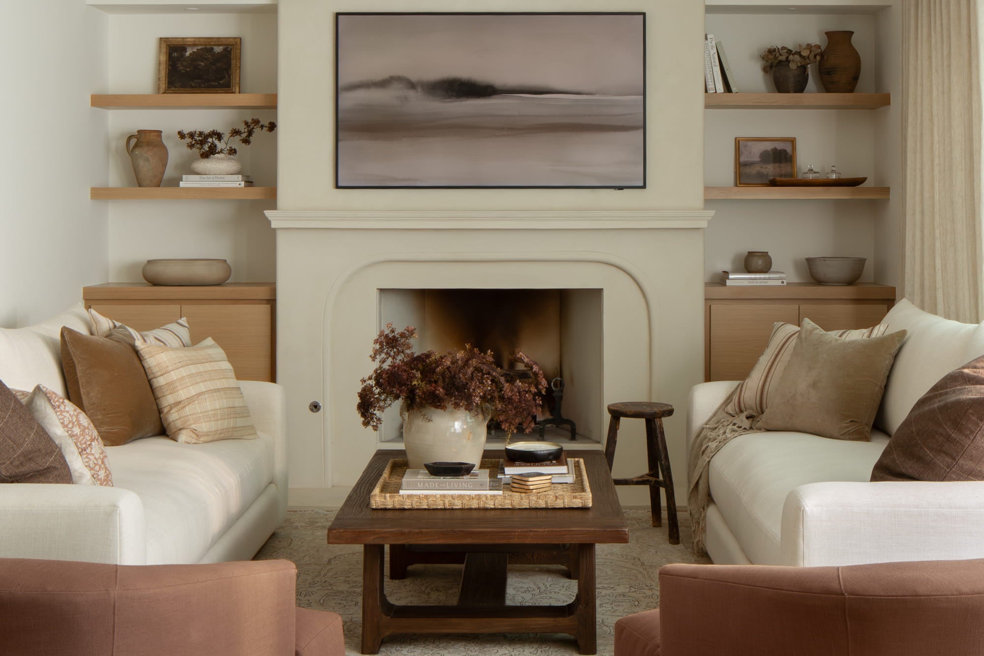 Symmetrical living room with plaster fireplace, floating wood shelves, and layered neutral pillows.