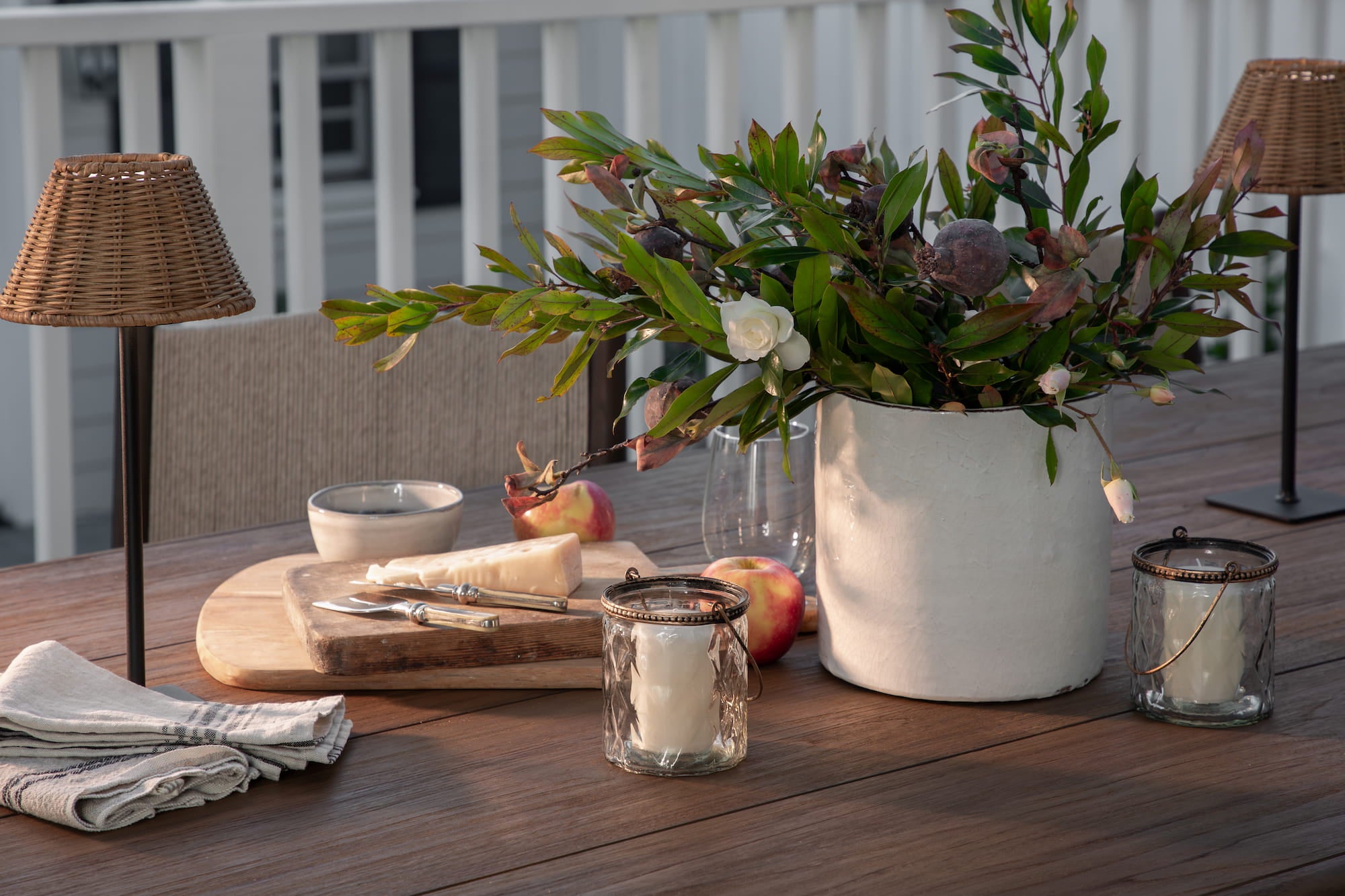 Outdoor wood dining table styled with cutting board, cheese knife, apples, glass candle holders, and greenery in white ceramic vase.