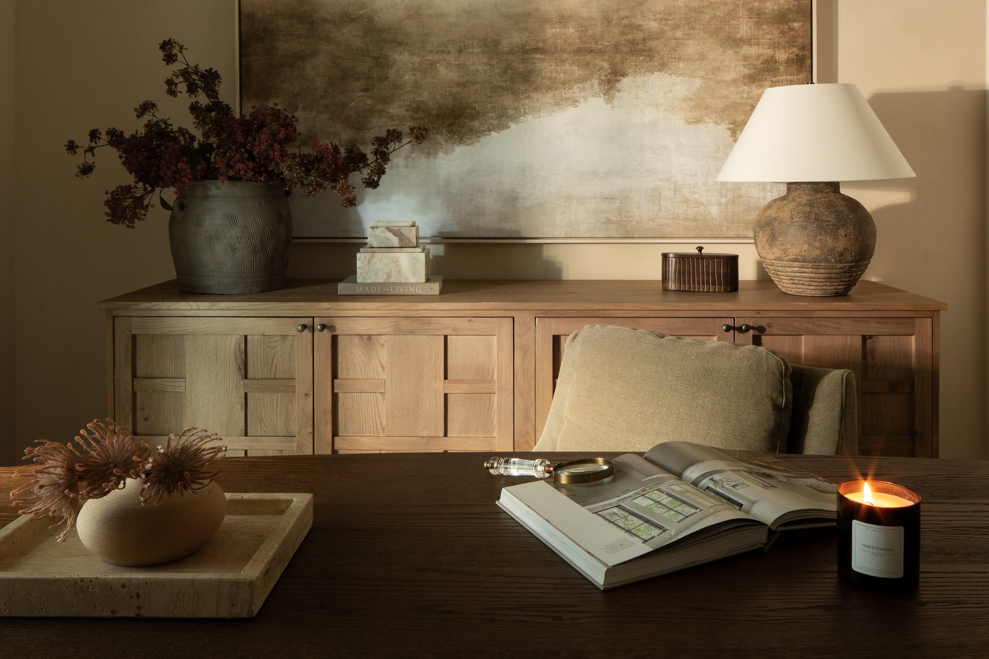 Wood desk styled with open design book and candle in front of textured artwork and wood credenza.