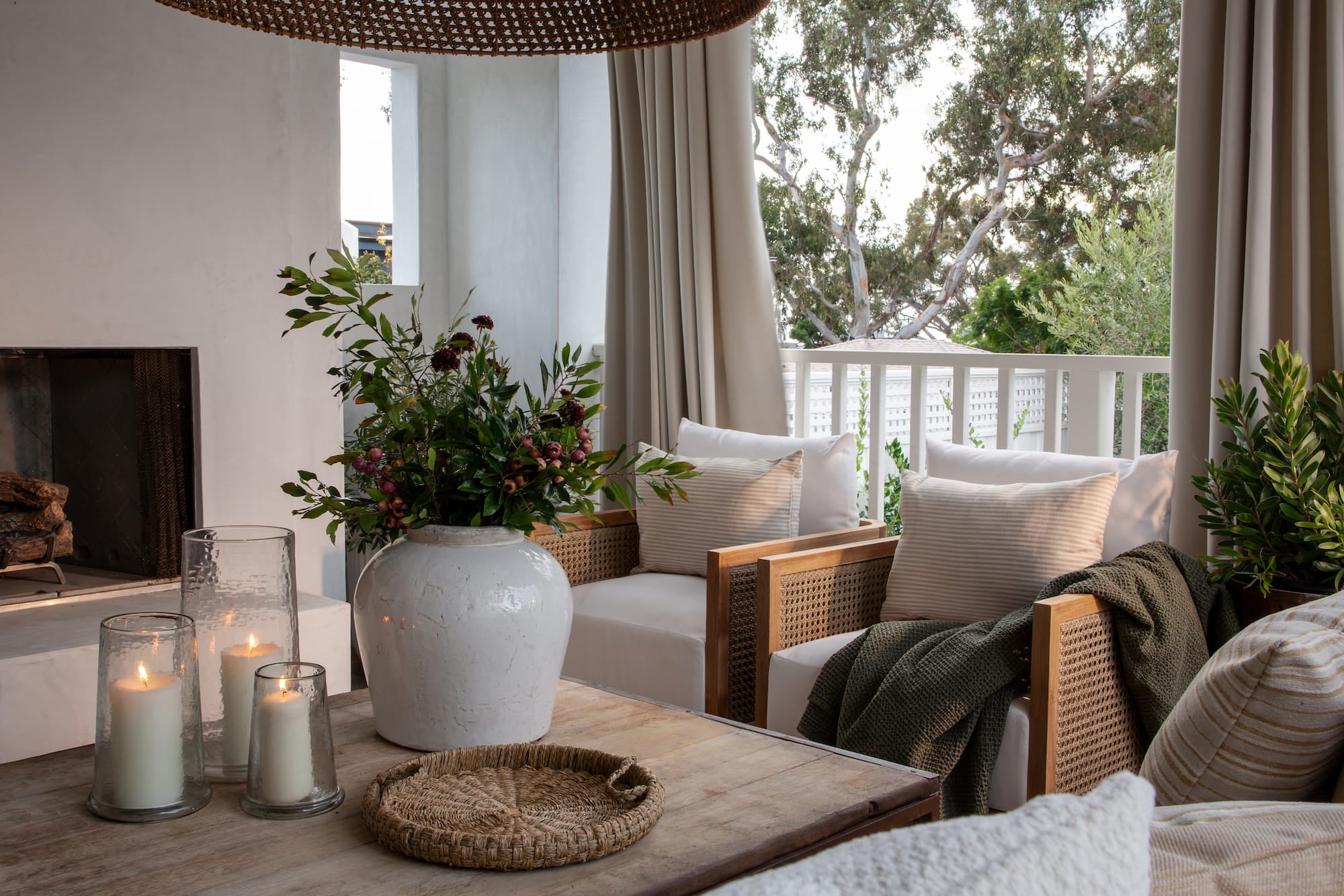 Covered outdoor patio with cushioned wood lounge chairs, woven coffee table, lit candles, and white railing overlooking trees.