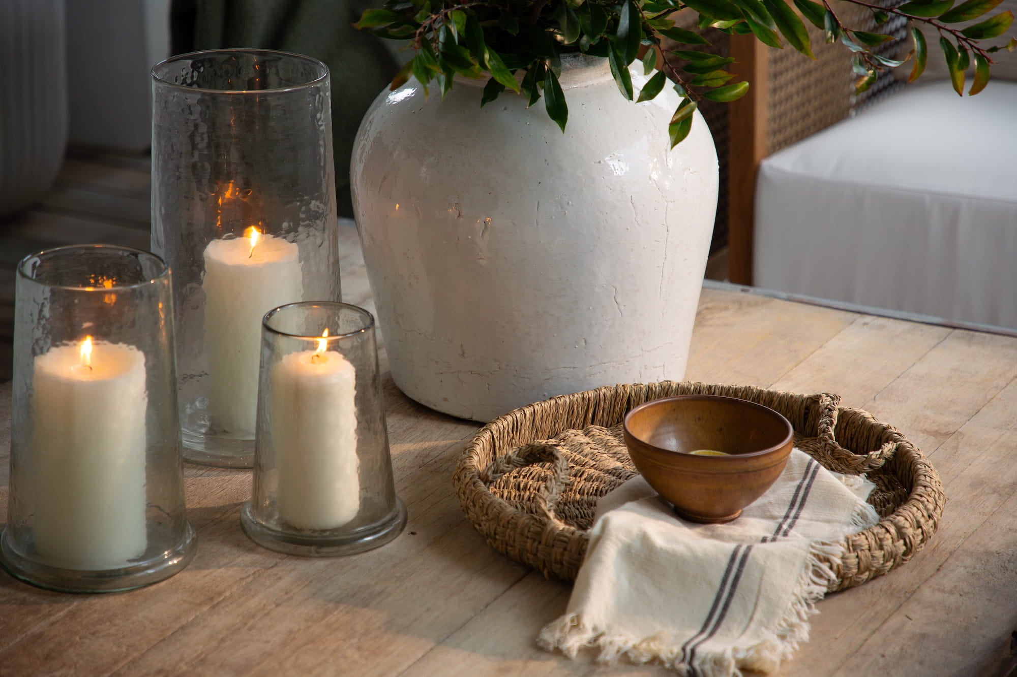 Wood coffee table styled with glass hurricane candles, woven tray, linen napkin, and white ceramic vase with greenery.