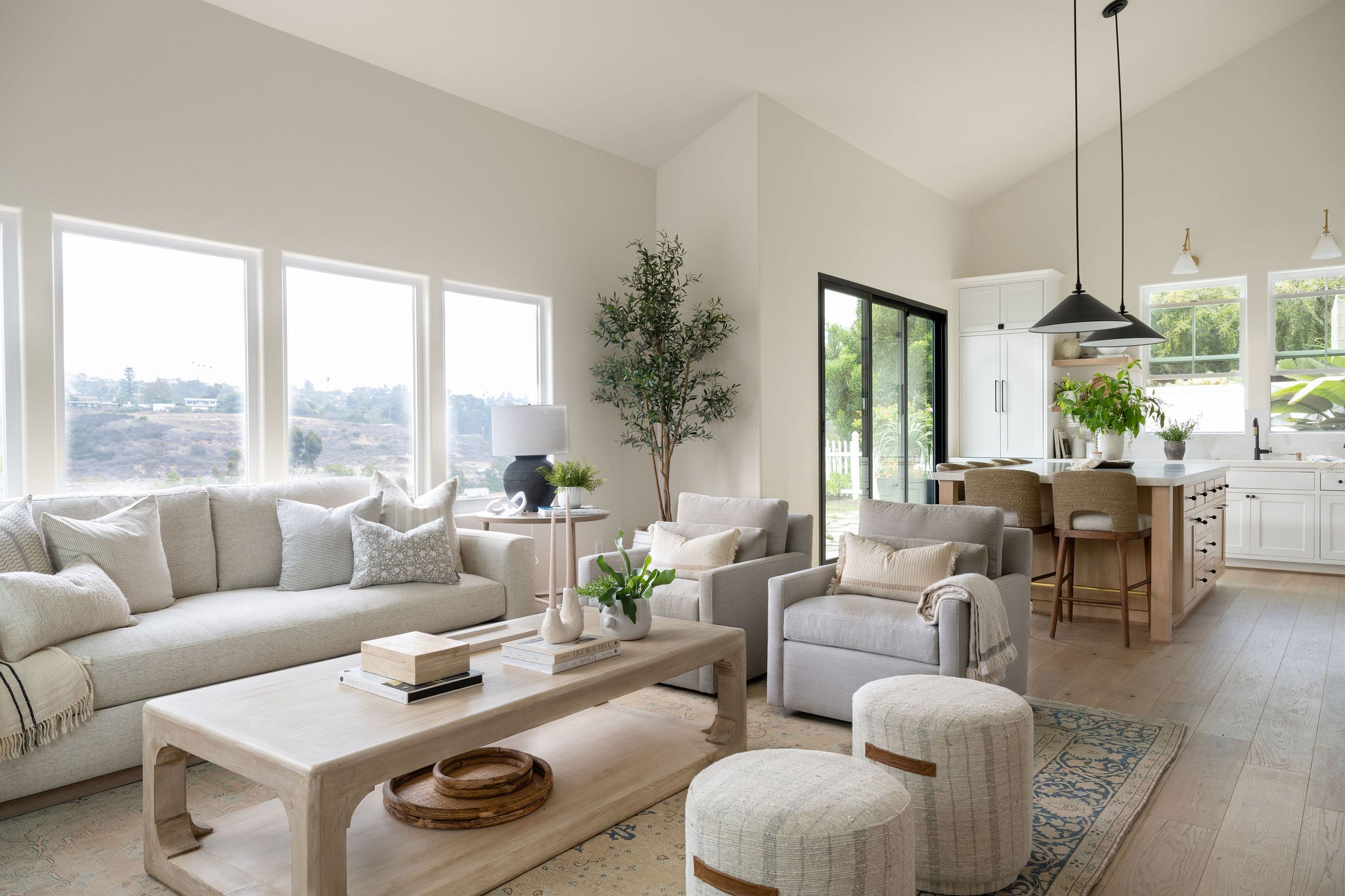 Modern living room with white furniture, large windows, and a kitchen in the background.