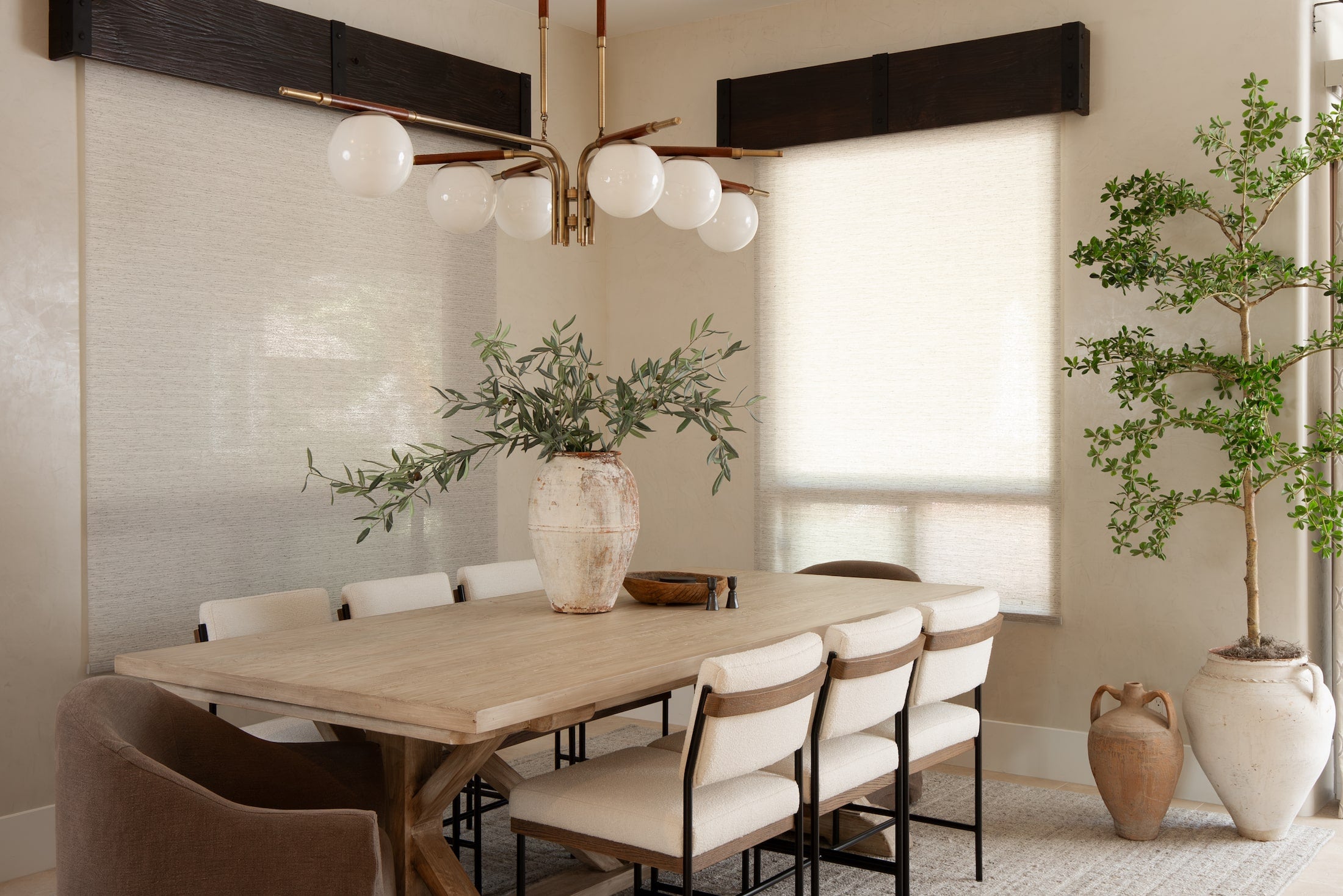 Dining room with light wood table, upholstered chairs, brass chandelier, and potted tree.