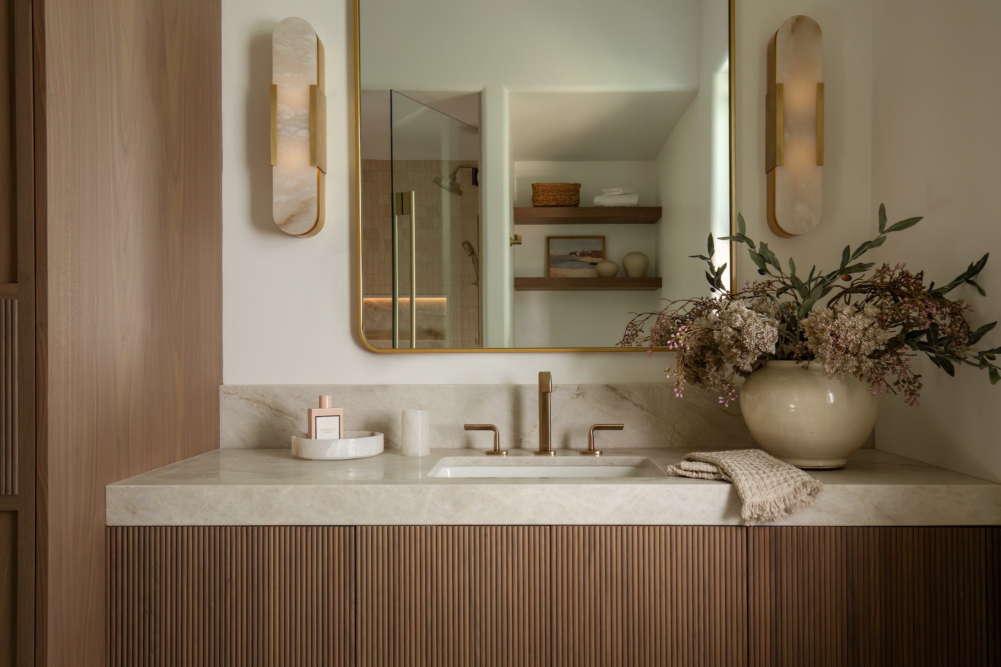Bathroom vanity with fluted wood cabinet, stone countertop, brass faucet, gold-framed mirror, and wall sconces.