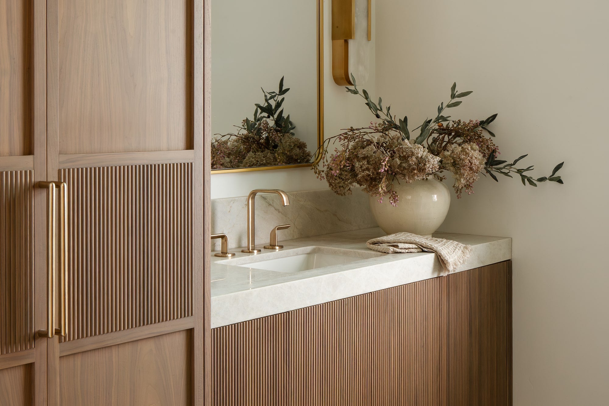 Bathroom with fluted wood vanity, stone countertop, brass faucet, gold-framed mirror, and tall wood wardrobe.