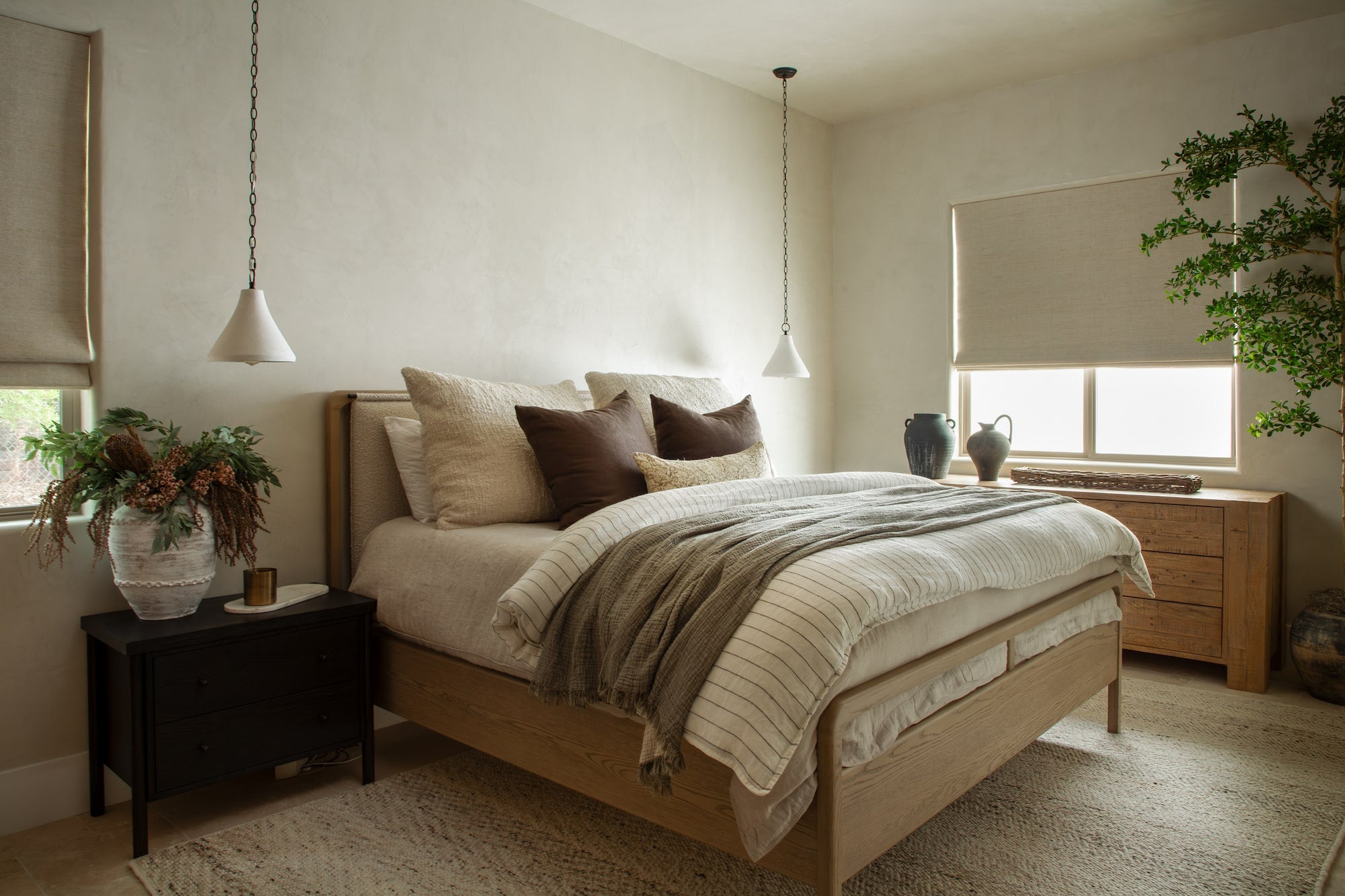 Bedroom with wood platform bed, layered neutral bedding, pendant bedside lights, and wood dresser beneath window.