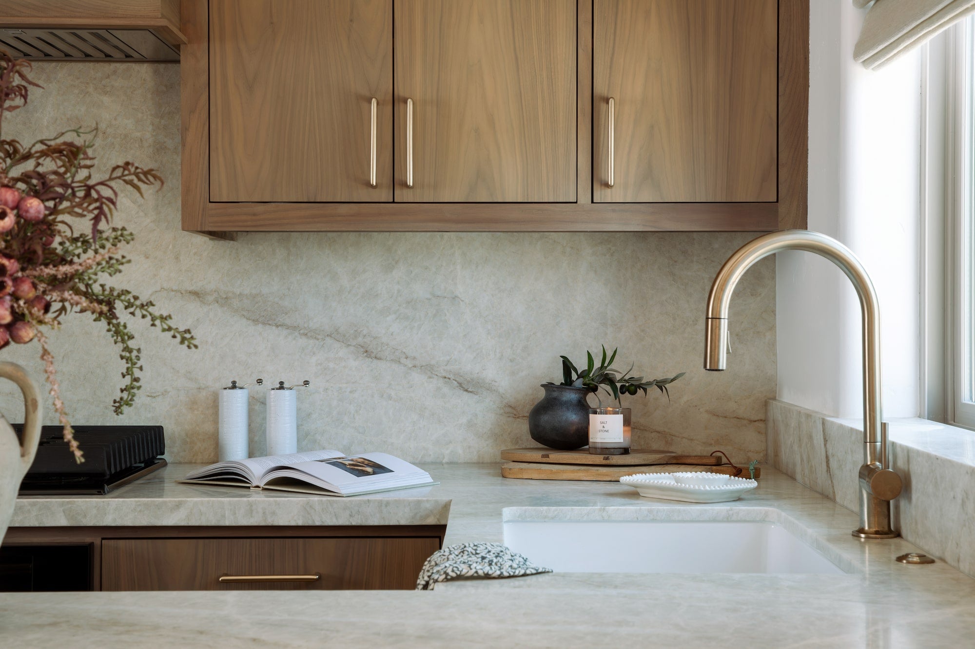 Wood kitchen cabinetry with brass pulls and stone backsplash surrounding cooktop and vent hood.
