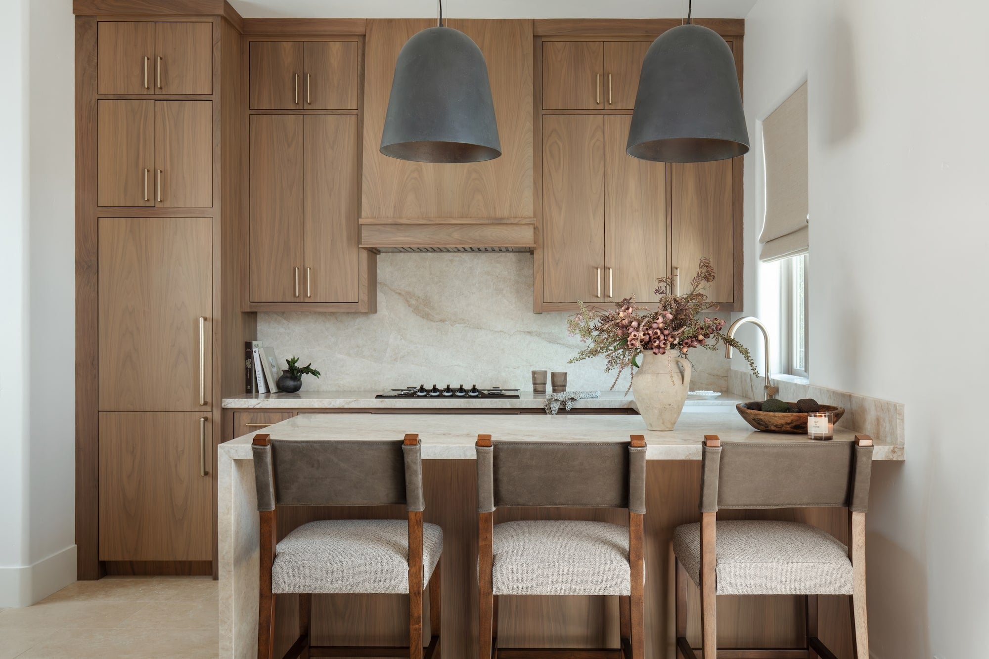 Kitchen with warm wood cabinetry, stone waterfall island, leather bar stools, and dark pendant lights overhead.
