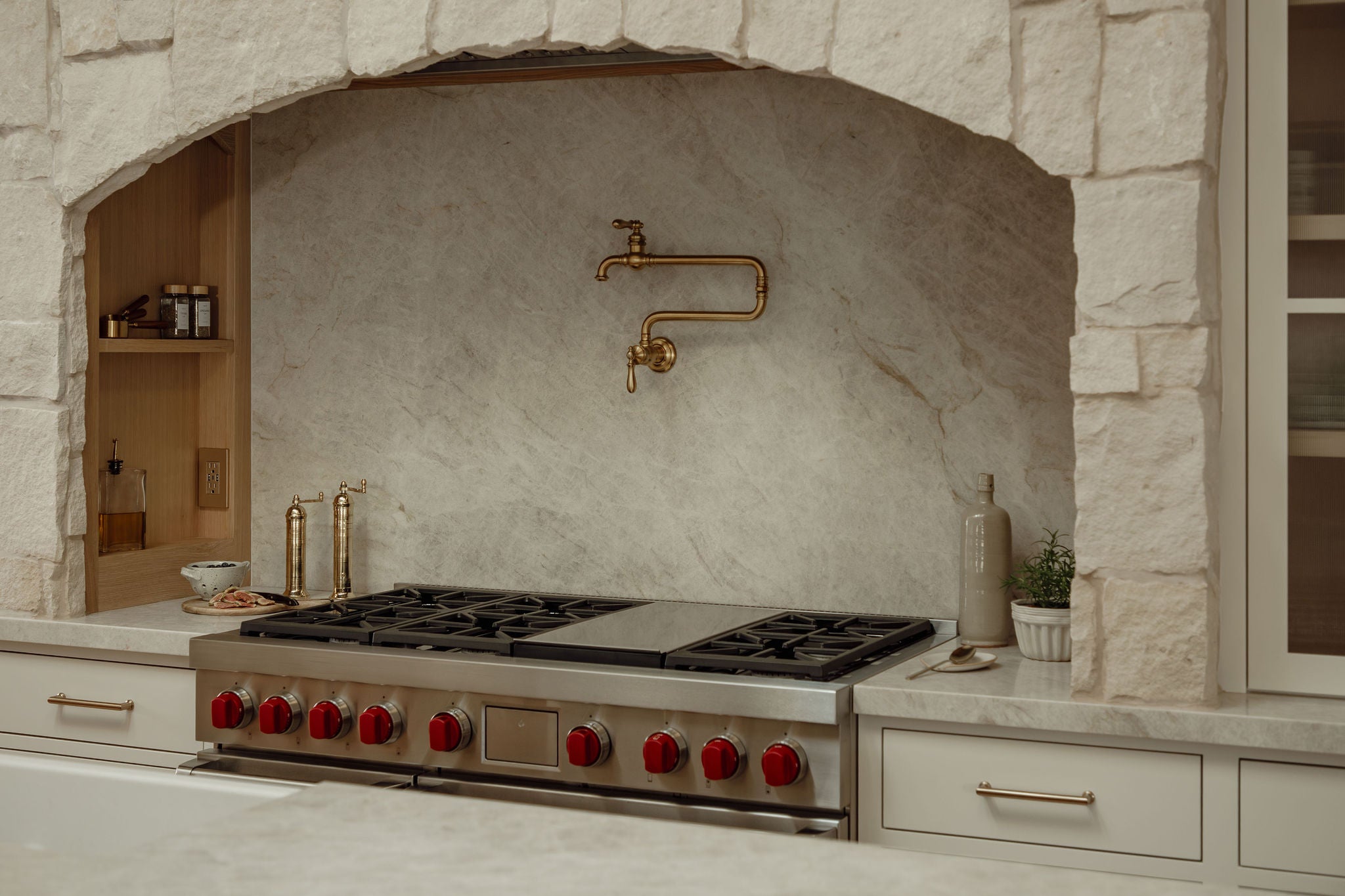 Professional gas range with red knobs beneath stone arch, marble backsplash, and brass pot filler.
