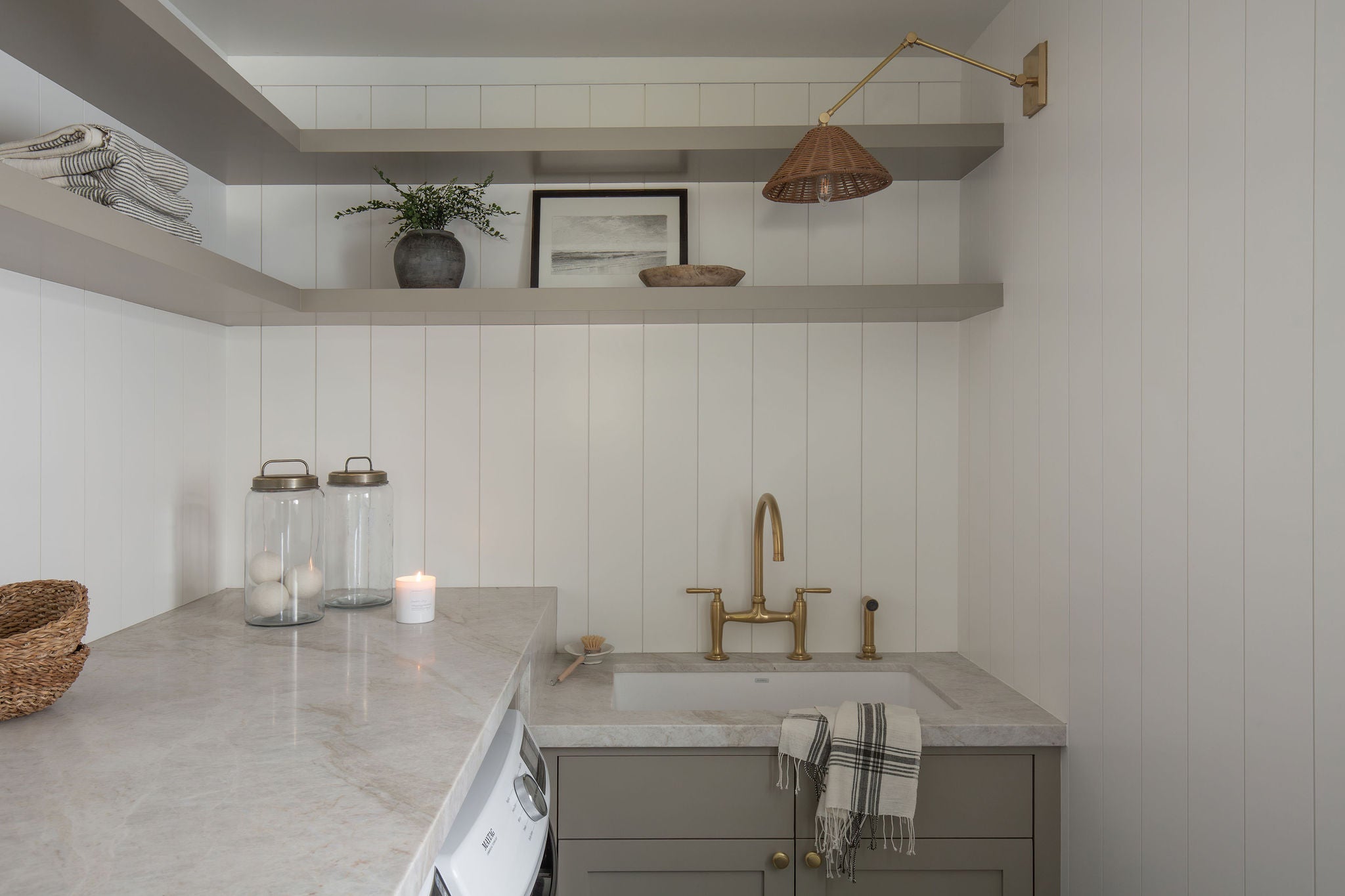 Laundry room with marble countertops, utility sink, brass faucet, floating shelves, and stacked washer beneath counter.